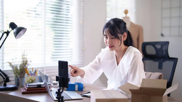 Young asian merchant women preparing smartphone to recording live streaming and selling clothes online in vlogger while working and shipping delivery for online shopping business in home office.