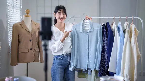 Young asian merchant women showing new fashion clothes to recording video and selling in social media on smartphone while working and shipping delivery for online shopping business in home office.