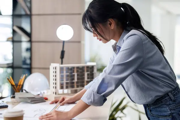 Asian architect engineer women using ruler to checking and drawing layout of architecture plan on blueprint while working to designing new project about building model and construction architectural.