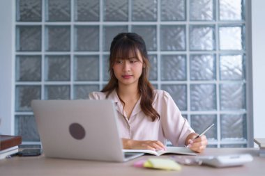 Gündelik giysiler içinde Asyalı genç bir öğrenci, üniversitede dizüstü bilgisayarlı dijital teknolojiyle çevrimiçi öğretmenlik eğitimini öğrenirken ve öğrenirken not defterine bilgi verileri yazıyor..