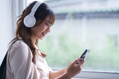 Genç Asyalı bir öğrenci, kulaklık takıyor, müzik dinliyor ve internette akıllı telefondan sörf yapıyor. Kitap okuduktan ve eğitim bilgisi öğrendikten sonra üniversitede ödev yapıyor..