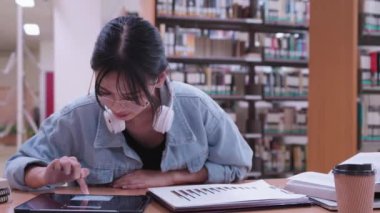 Genç Asyalı üniversite öğrencileri, üniversite kütüphanesinde kulaklık takarak tabletlerinde müzik dinleyerek çalışmaktan keyif alıyorlar. Yüksek kalite 4k görüntü