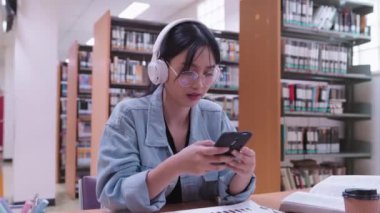 Genç Asyalı kız üniversite öğrencileri, üniversite kütüphanesinde kulaklık takarak, akıllı telefonlarıyla müzik dinleyerek çalışmaktan keyif alıyorlar. Yüksek kalite 4k görüntü
