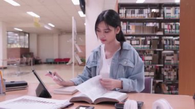Asyalı bir kız öğrenci üniversite kütüphanesinde tabletinin içeriğini okumaya ve özetlemeye daldı. Yüksek kalite 4k görüntü