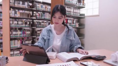 Asyalı bir kız öğrenci üniversite kütüphanesinde tabletinin içeriğini okumaya ve özetlemeye daldı. Yüksek kalite 4k görüntü
