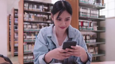 Asyalı kız öğrenci üniversite kütüphanesinde akıllı telefonuyla sosyal medyaya bağlanıyor. Yüksek kalite 4k görüntü
