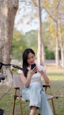 Asyalı kadın halka açık parkta bisiklet sürmeye ara verirken telefonda vakit geçirmekten hoşlanıyor. Doğanın ortasında teknolojiyle rahatlamak. Yüksek kalite 4k görüntü