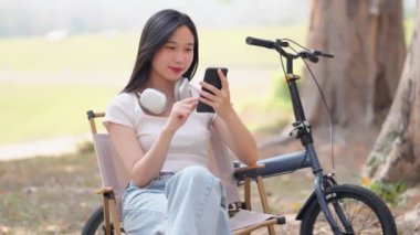 Asyalı kadın halka açık parkta bisiklet sürmeye ara verirken telefonda vakit geçirmekten hoşlanıyor. Doğanın ortasında teknolojiyle rahatlamak. Yüksek kalite 4k görüntü