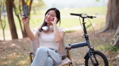 Asyalı kadın cep telefonuyla selfie çekiyor, parka bisikletle gitmeye ara veriyor. Doğanın içinde boş zamanların tadını çıkarıyorum. - Evet. Yüksek kalite 4k görüntü