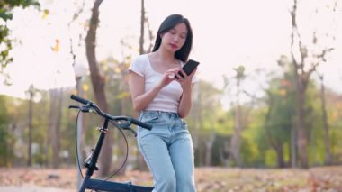 Parkta bisiklet sürdükten sonra telefonuyla dinlenen Asyalı bir kadın. Huzurlu anların tadını çıkarmak ve doğanın güzelliğini yakalamak. - Evet. Yüksek kalite 4k görüntü