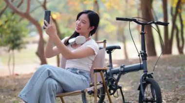 Asyalı kadın cep telefonuyla selfie çekiyor, parka bisikletle gitmeye ara veriyor. Doğanın içinde boş zamanların tadını çıkarıyorum. - Evet. Yüksek kalite 4k görüntü