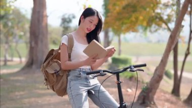 Asyalı kadın halk parkında kitap okuyup bisikletçiliğe ara vermenin tadını çıkarıyor. Doğanın ortasında iyi bir okumayla rahatlıyorum. Yüksek kalite 4k görüntü