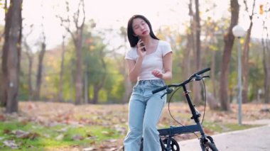 Parkta bisiklet sürdükten sonra telefonuyla dinlenen Asyalı bir kadın. Huzurlu anların tadını çıkarmak ve doğanın güzelliğini yakalamak. - Evet. Yüksek kalite 4k görüntü