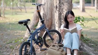Asyalı kadın halk parkında kitap okuyup bisikletçiliğe ara vermenin tadını çıkarıyor. Doğanın ortasında iyi bir okumayla rahatlıyorum. Yüksek kalite 4k görüntü