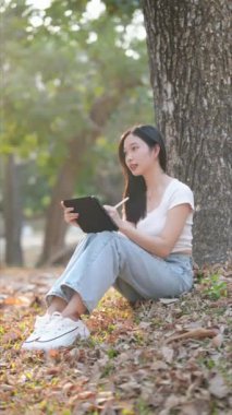 Kamu parkında barışçıl bir mola veren Asyalı kadın tablet çizimleriyle yaratıcılığını ifade ediyor. Doğanın ortasında rahatlatıcı anlar. Yüksek kalite 4k görüntü