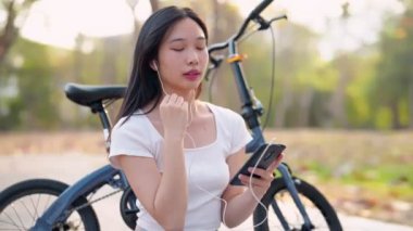 Asyalı kadın en sevdiği şarkıları seçmek ve dinlemek için cep telefonunu kullanıyor, parktaki bisikletçiliğe ara veriyor. Doğanın ortasındaki müziğin tadını çıkarıyorum. Yüksek kalite 4k görüntü