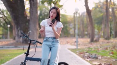 Parkta bisiklet sürdükten sonra telefonuyla dinlenen Asyalı bir kadın. Huzurlu anların tadını çıkarmak ve doğanın güzelliğini yakalamak. - Evet. Yüksek kalite 4k görüntü