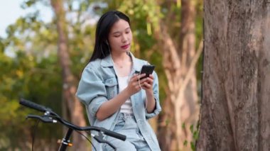 Asyalı kadın en sevdiği şarkıları seçmek ve dinlemek için cep telefonunu kullanıyor, parktaki bisikletçiliğe ara veriyor. Doğanın ortasındaki müziğin tadını çıkarıyorum. Yüksek kalite 4k görüntü