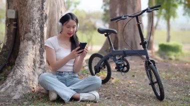 Asyalı kadın cep telefonu kullanıyor favori şarkıları dinliyor, bisiklete ara veriyor. Yüksek kalite 4k görüntü