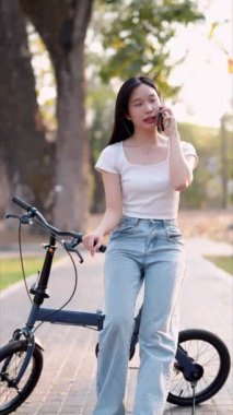 Parkta bisiklet sürdükten sonra telefonuyla dinlenen Asyalı bir kadın. Huzurlu anların tadını çıkarmak ve doğanın güzelliğini yakalamak. - Evet. Yüksek kalite 4k görüntü