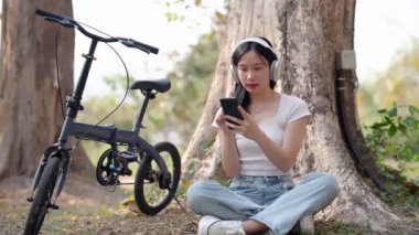 Asyalı kadın cep telefonu kullanıyor favori şarkıları dinliyor, bisiklete ara veriyor. Yüksek kalite 4k görüntü