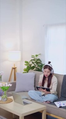 Evden çalışan Asyalı iş kadını tabletine notlar yazıyor ve oturma odasındaki kanepede dizüstü bilgisayarı üzerinde çalışıyor. Yüksek kalite 4k görüntü