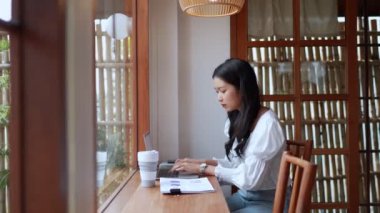 Asyalı genç bir iş kadını, yakınlardaki bir kafede bilgisayarını kullanarak çalışıyor. Yüksek kalite 4k görüntü