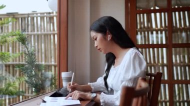 Yakındaki bir kafede çalışan Asyalı bir bayan girişimci tablet kullanarak görevlerini özenle not alıyor ve odaklanmış çalışmalarını gösteriyor. Yüksek kalite 4k görüntü