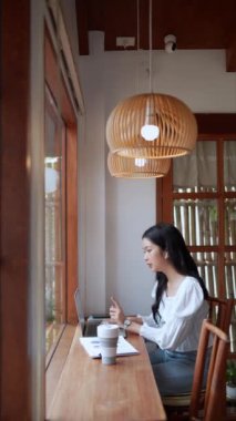 Yakındaki bir kafede çalışan Asyalı bir bayan girişimci, meslektaşlarıyla kablosuz kulaklık kullanarak konuşuyor, bilgisayarına odaklanıyor, kendini adamış ve kararlı olduğunu gösteriyor. Yüksek kalite 4k görüntü
