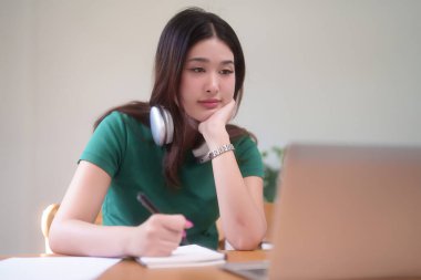 Boynuna kulaklık takan Asyalı genç bir öğrenci ve çevrimiçi derste internetteki web sitelerini izleyip evden bilgi öğrenirken ve öğrenirken not defterine bilgi yazıyorlar..