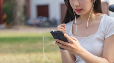 Genç Asyalı kadınların kulaklık takıp akıllı telefondan müzik dinlemeleri ve eğlence izlemeleri parkta bisiklet sürdükten sonra seyahat yaşam tarzıyla rahatlamalarıdır..