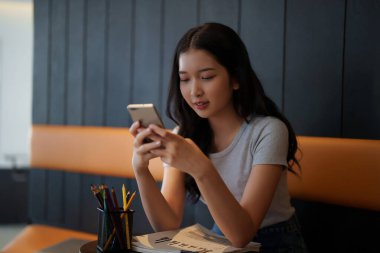 Asyalı genç bayan, kafeteryadaki eğitim bilgisi hakkında bilgi edinmek için okuduktan sonra arkadaşlarıyla sohbet etmek ve sosyal medyada sörf yapmak için akıllı telefon kullanıyor..