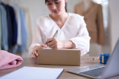 Genç Asyalı tüccar kadınlar karton kutuya adres verileri yazıyorlar ve online siparişleri kontrol ederken teslimat müşterilerini göndermeye hazırlanıyorlar ve çevrimiçi alışveriş işleri hakkında çalışıyorlar..