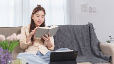 Asyalı iş kadını evde teknoloji tabletleriyle çalışırken pazarlama stratejisi ve yeni iş projelerinin yatırımı hakkında plan yaptıktan sonra koltukta oturup kitap okuyarak rahatlıyor..