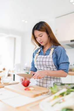 Genç Asyalı bir kadın bıçakla tahtayı keserken elma dilimlerken kâsede taze sebze salatası ve evde sağlıklı yaşam tarzıyla modern mutfakta sağlıklı kahvaltı hazırlıyor..