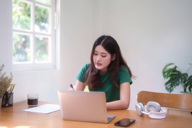 Genç Asyalı kadın öğrenciler ders çalışırken ve izlerken dizüstü bilgisayarda ödev yaparken aynı zamanda not defterine bilgi yazıp evden bilgi uzaktan eğitimi alıyorlar..