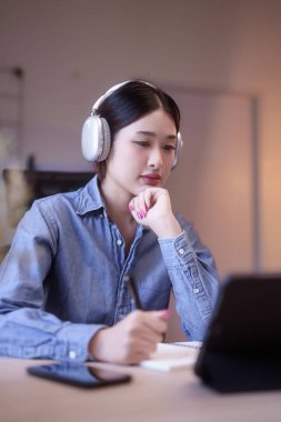 Genç Asyalı kadın öğrencilerin kulaklık takıp, online derste, tablet üzerinde, eğitim hakkında düşünceli bir şekilde notlar yazmayı ve evden bilgi uzaktan eğitimi almayı öğrenmeleri..
