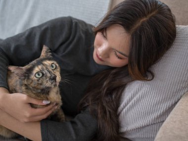 Kanepede uzanmış, kaplumbağa kabuğu kedisine sevgiyle sarılan bir kadın. Samimi an, güçlü bağı ve rahatlığı yansıtır. 