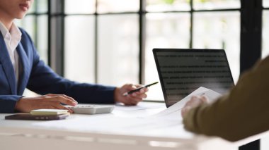  Masada çalışan iki kişinin olduğu profesyonel bir ortam. Belgeleri incelemekle ve hesap makinesi kullanmakla meşguller. Dizüstü bilgisayarlar masada açık.