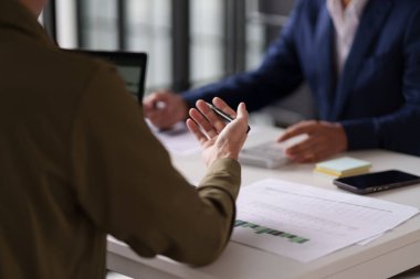  Masada çalışan iki kişinin olduğu profesyonel bir ortam. Belgeleri incelemekle ve hesap makinesi kullanmakla meşguller. Dizüstü bilgisayarlar masada açık.