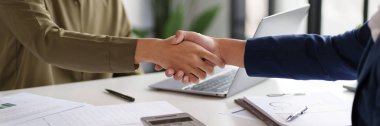 İki kişi masada el sıkışıyor, bu da başarılı bir anlaşma ya da ortaklık olduğunu gösteriyor. Masa belgelerle dolu.