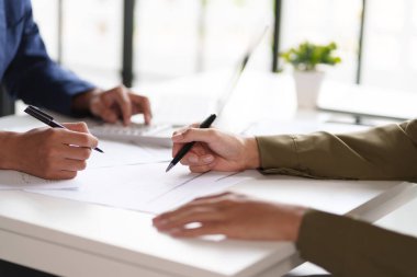 Masadaki bir belgeyi tartışan ve işaret eden iki kişinin yakın çekimi. Elleri kalemleri tutuyor, belgenin içeriğine ilişkin bir inceleme veya işbirliği gösteriyor.