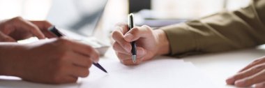 Masadaki bir belgeyi tartışan ve işaret eden iki kişinin yakın çekimi. Elleri kalemleri tutuyor, belgenin içeriğine ilişkin bir inceleme veya işbirliği gösteriyor.