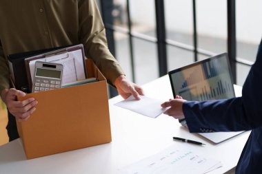 İki kişi bir masada belgeleri değiş tokuş ediyor. Bir kişi ofis malzemeleriyle dolu bir kutu tutuyor. Bu da olası bir iş değişimi ya da proje devrini gösteriyor..