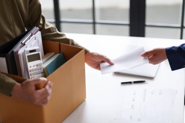 İki kişi bir masada belgeleri değiş tokuş ediyor. Bir kişi ofis malzemeleriyle dolu bir kutu tutuyor. Bu da olası bir iş değişimi ya da proje devrini gösteriyor..