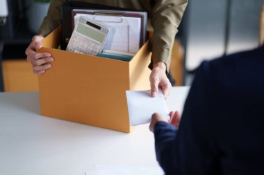 İki kişi bir masada belgeleri değiş tokuş ediyor. Bir kişi ofis malzemeleriyle dolu bir kutu tutuyor. Bu da olası bir iş değişimi ya da proje devrini gösteriyor..