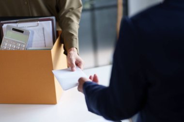 İki kişi bir masada belgeleri değiş tokuş ediyor. Bir kişi ofis malzemeleriyle dolu bir kutu tutuyor. Bu da olası bir iş değişimi ya da proje devrini gösteriyor..