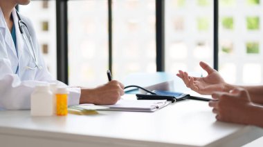 Muayene sırasında bir doktor ve hasta. Hasta semptomlarını açıklarken doktor panoya yazı yazıyor. Masada tıbbi şişeler ve kan basıncı monitörü var..
