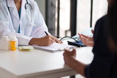 Muayene sırasında bir doktor ve hasta. Hasta semptomlarını açıklarken doktor panoya yazı yazıyor. Masada tıbbi şişeler ve kan basıncı monitörü var..