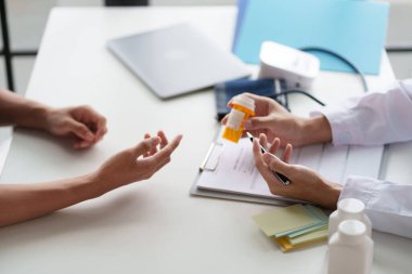 Bir hastaya ilacı açıklayan bir doktor. Doktor elinde reçeteli bir şişe tutuyor ve bir panoya yazı yazıyor. Tıbbi şişeler masada, tedavi hakkında detaylı bir tartışmayı gösteriyor..